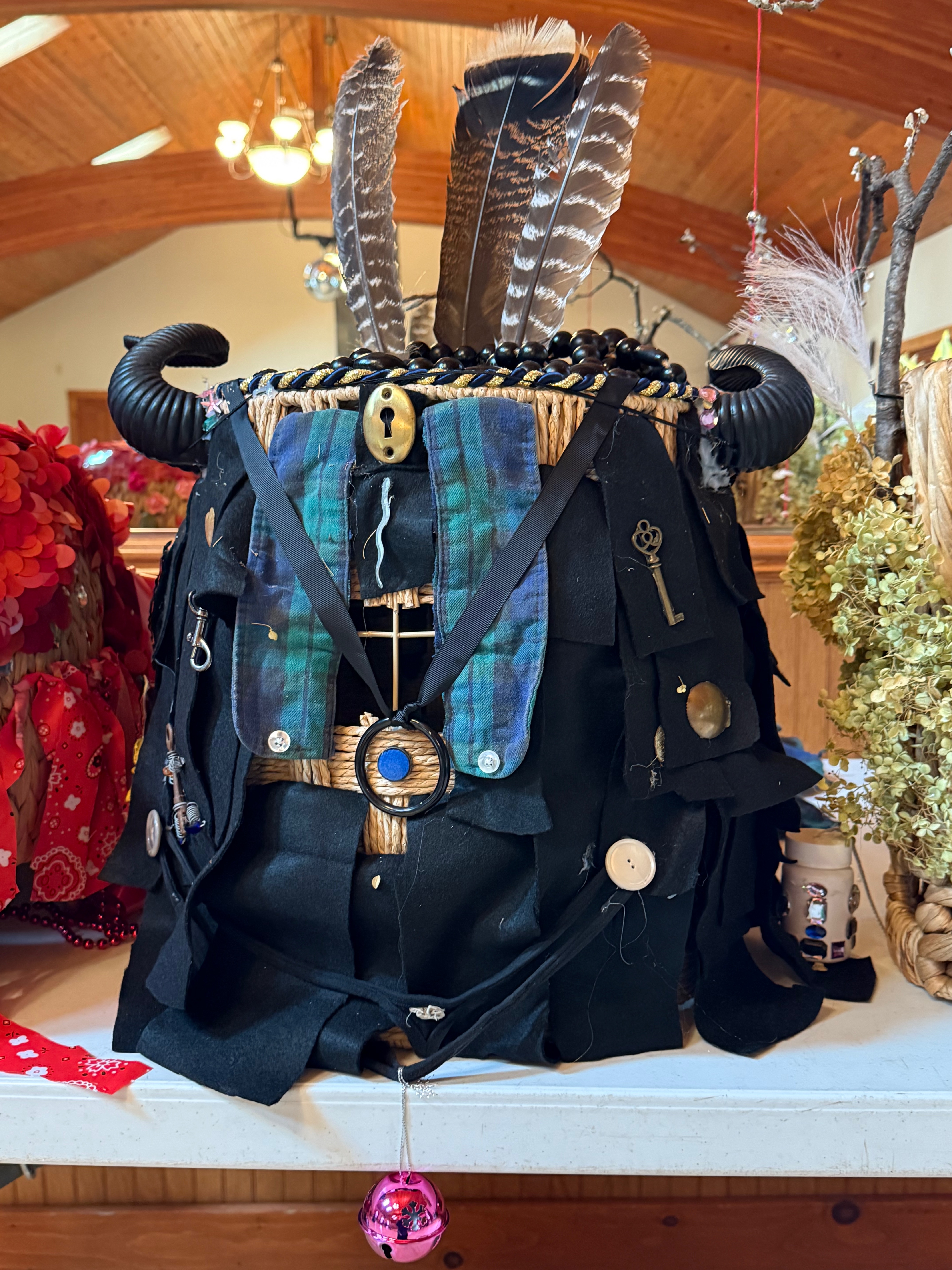 A headdress made from strips of black felt folded into a woven basket with an eyehole cut out. Baubles are glued to the black felt including a key, a keyhole, buttons, and a snake pin. A black cock ring dangles just below the eye hole. Black horns flank the crown which is topped with turkey feathers.