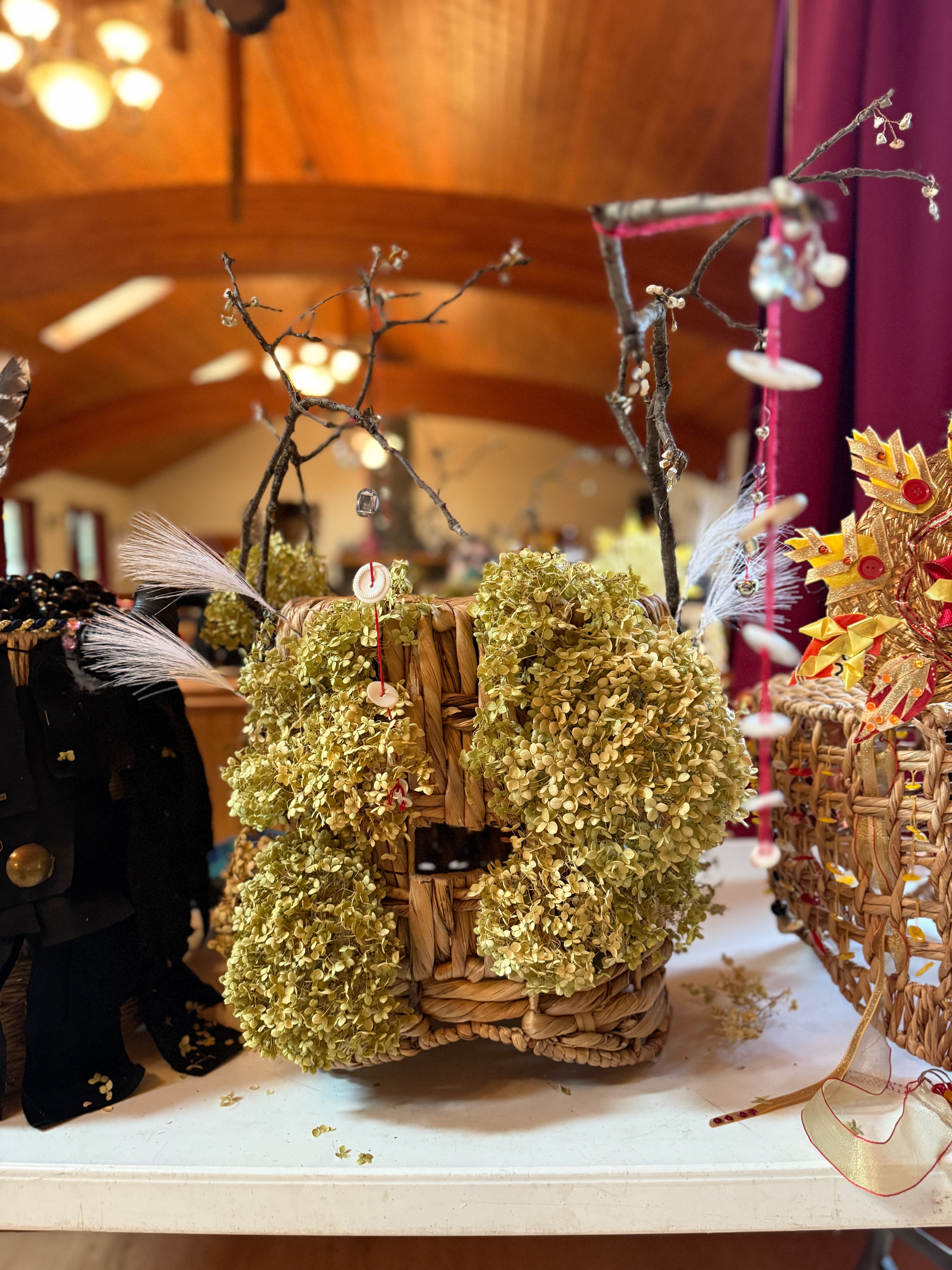 A headdress made from dried white hydrangea blossoms atttached to a woven basket. Sticks protude from the crown like antlers. Buttons are suspended with red thread from various tips of the sticks.