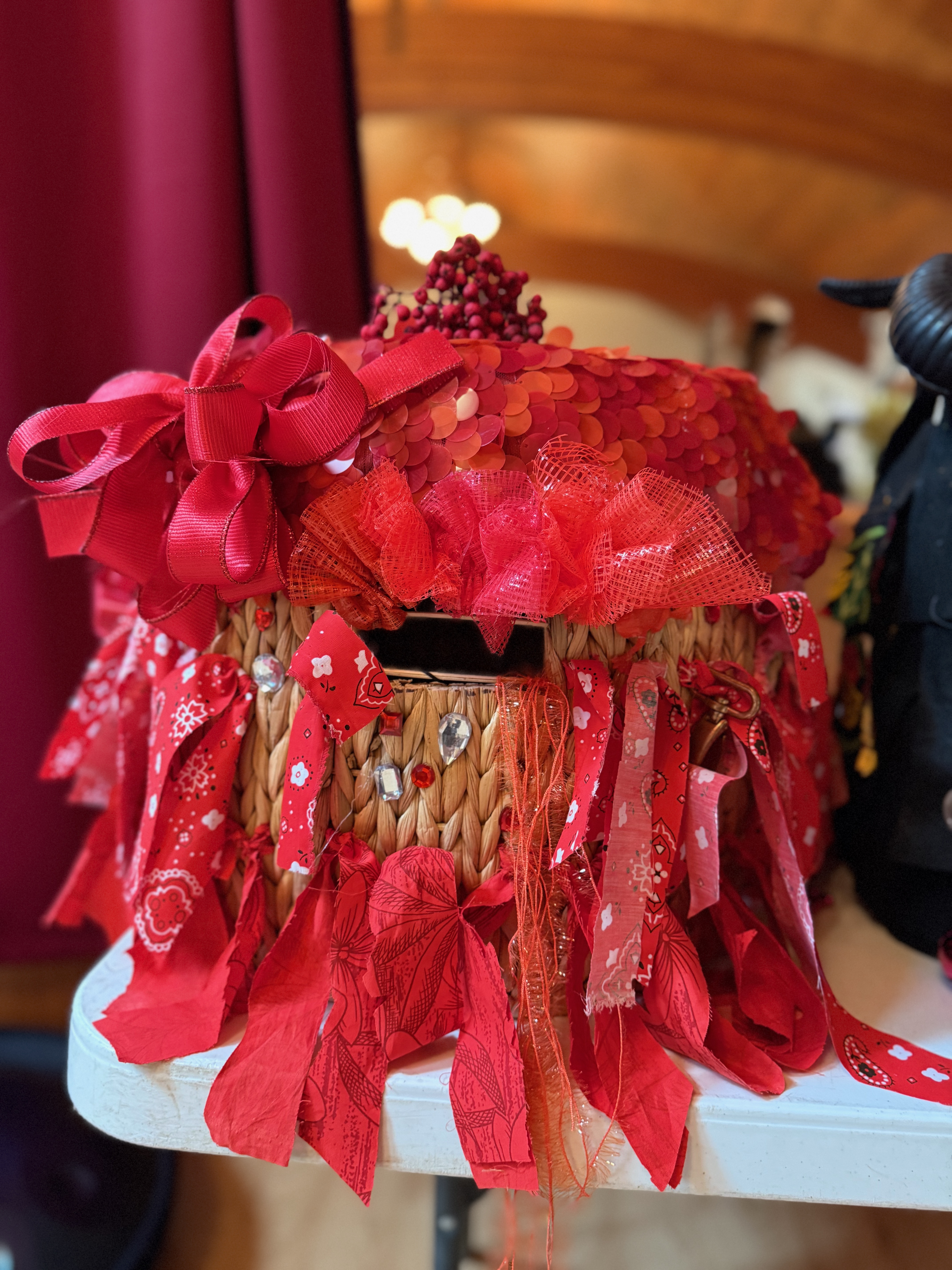 A headdress made from strips of red fabric threaded into a woven basket with an eyehole cut out. There are gems placed here and there between the fabric strips. The top is covered in a red sequined fabric and crowned with Toyon (Christmas berry).
