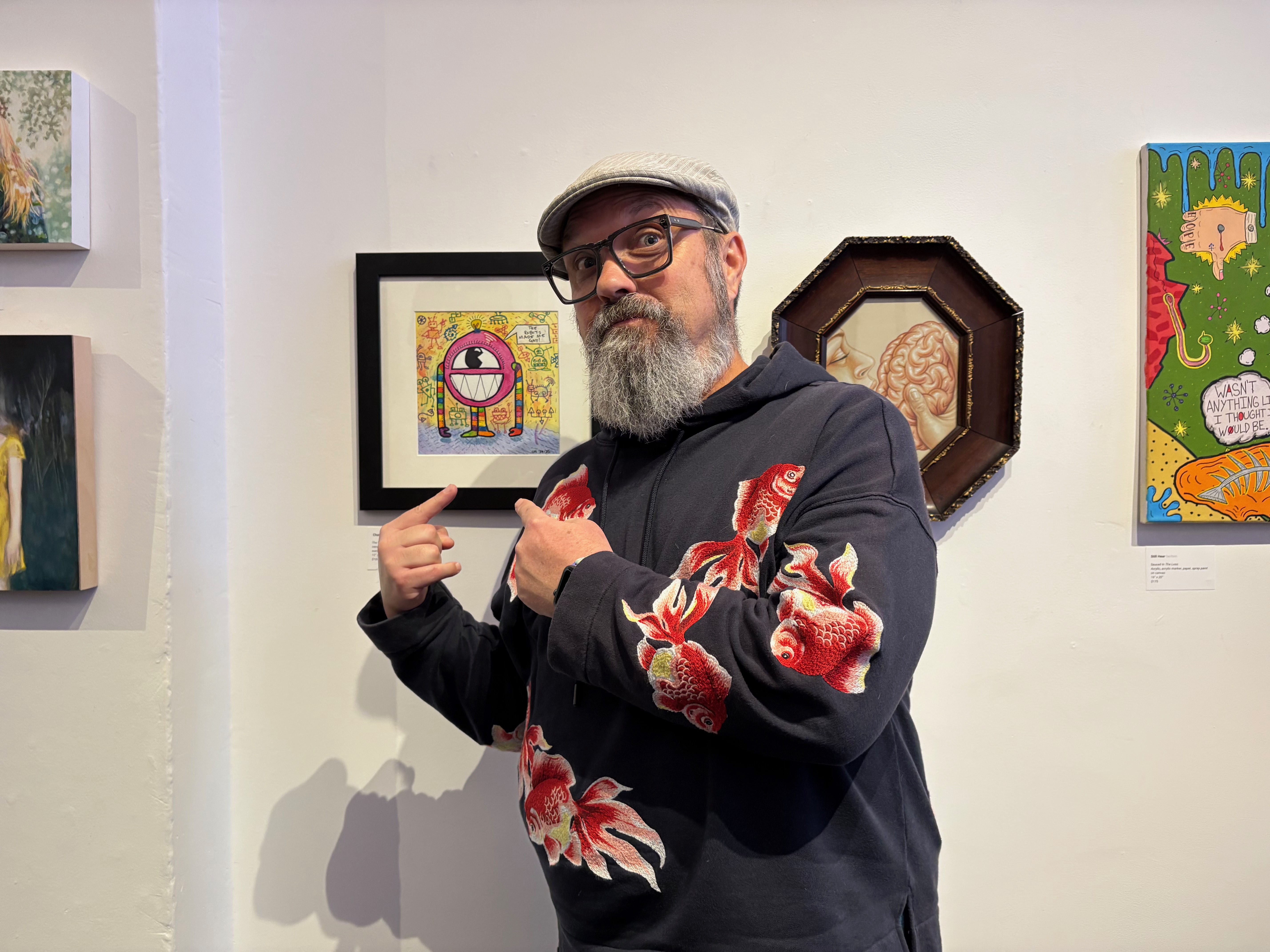 The bearded artist wearing a hoodie covered in embroidered goldfish points at his robot artwork hanging on the wall.