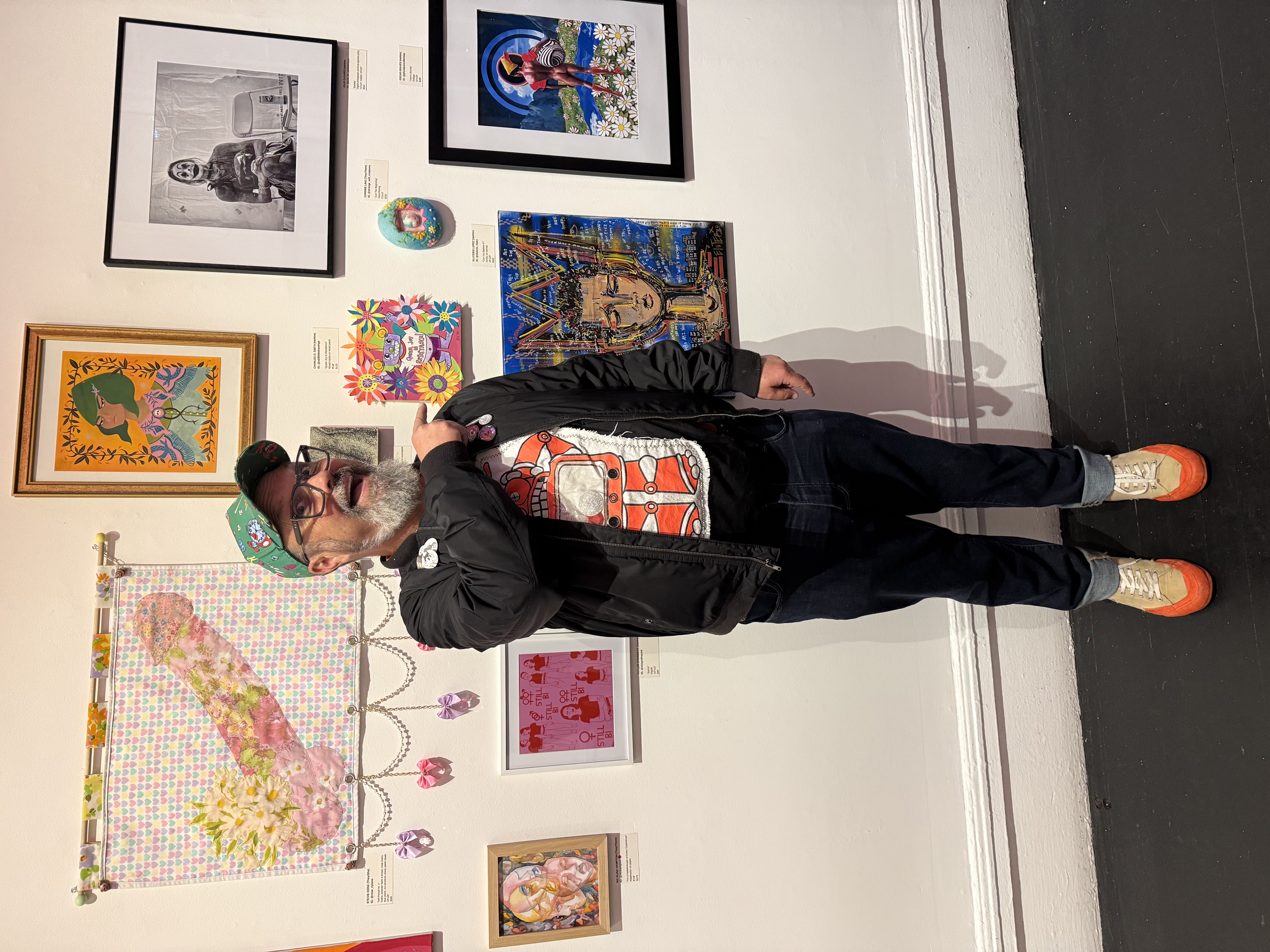 The artist points at his robot artwork surrouded by all the other art on the gallery wall.