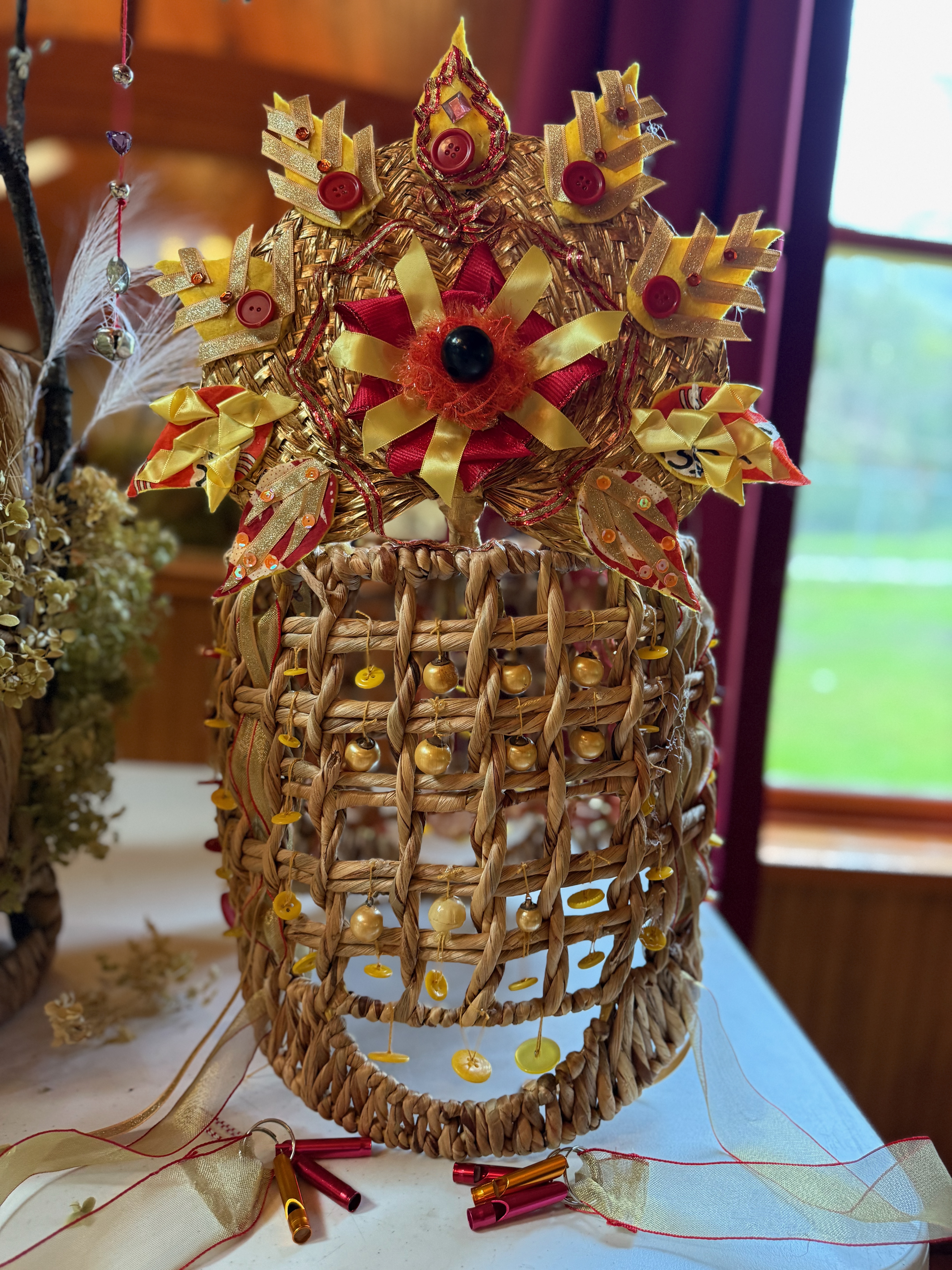 A headdress made from a woven basket. In the spaces between the basket weave, are suspended buttons on strings. An intricately detailed golden crown of flames made from felt, fabric, buttons, ribbon, and sequins adorns the top.