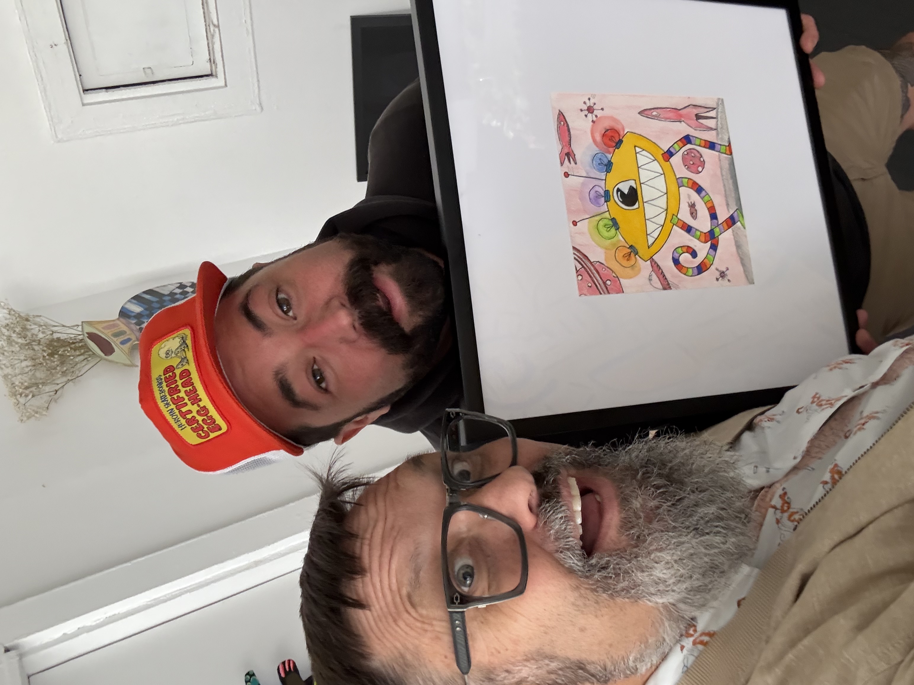 Cabure Bonugli in a red cap holding my framed art: Pink Planet. The drawing is of a lemon-shaped robot a space-scape of planets, rockets, and shooting stars.
